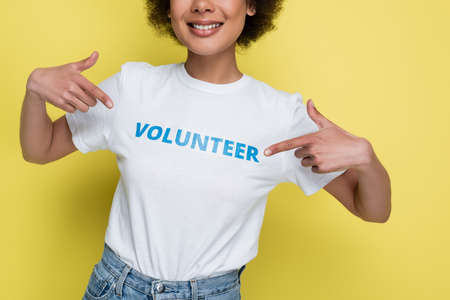 cropped view of smiling african american woman pointing at volunteer inscription on t-shirt isolated on yellowの写真素材