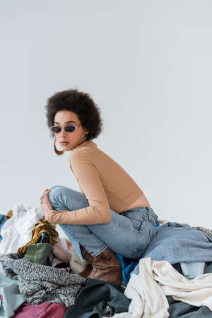 brunette african american woman in trendy sunglasses near pile of different clothes on grey backgroundの写真素材