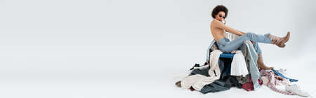 full length of african american woman sitting near pile of clothing on grey background, banner