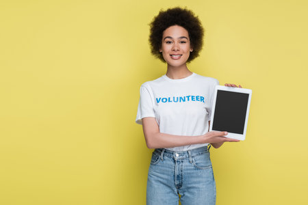 pretty african american volunteer showing digital tablet with blank screen isolated on yellow