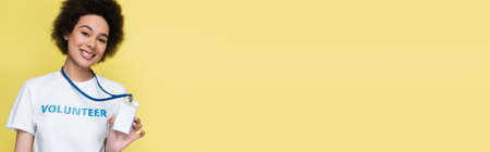 happy african american volunteer showing empty name tag at camera isolated on yellow, bannerの写真素材