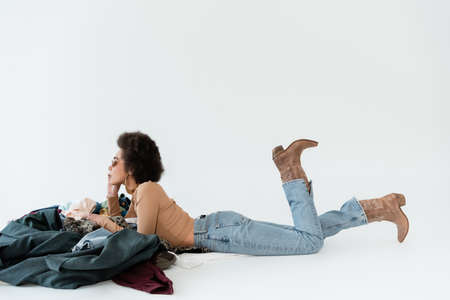 side view of african american woman in jeans and cowboy boots lying near pile of clothing on grey...