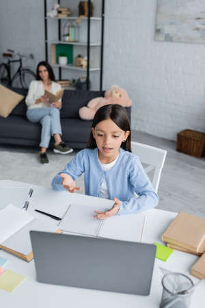 girl talking at laptop during online lesson near babysitter on blurred background