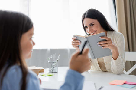 cheerful nanny with textbook near girl pointing with finger on blurred foreground