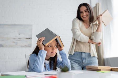bored girl covering head with textbook near nanny smiling on blurred backgroundの写真素材