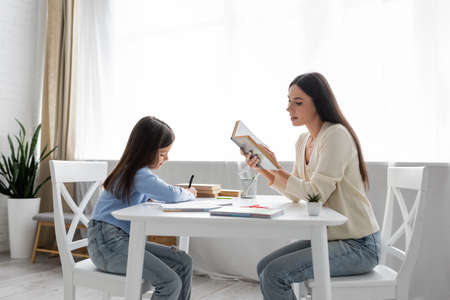side view of babysitter reading aloud near girl writing dictation at homeの写真素材