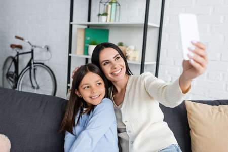 smiling babysitter taking selfie on cellphone with girl on sofa at home