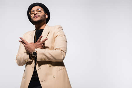 low angle view of indian hip hop performer in fedora hat and eyeglasses gesturing isolated on grey