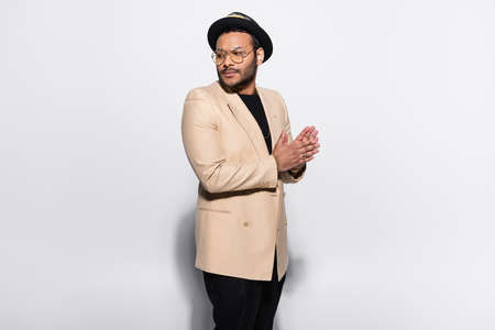 stylish indian hip hop performer in fedora hat and eyeglasses standing and looking away isolated on...