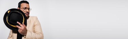 eastern hip hop performer in eyeglasses holding stylish fedora hat isolated on grey, bannerの写真素材