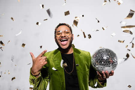 amazed indian hip hop performer in green velvet blazer and crown holding disco ball near falling...
