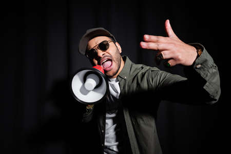 emotional middle east hip hop performer in sunglasses and cap singing and holding loudspeaker on blackの写真素材