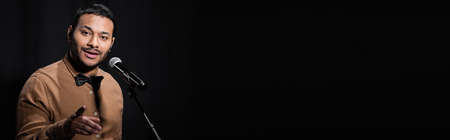 indian comedian telling jokes into microphone and pointing with finger while looking at camera isolated on black, bannerの写真素材