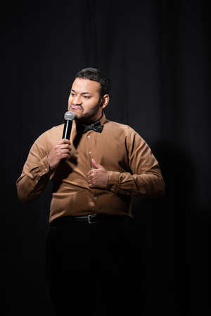indian stand up comedian grimacing and showing thumb up while telling jokes into microphone on blackの写真素材