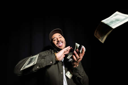 excited and rich indian hip hop performer in cap throwing dollar banknotes on black