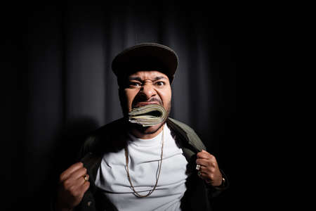 indian hip hop performer in cap biting bundle of dollar banknotes on black
