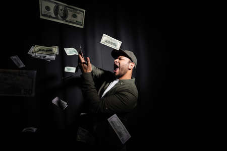excited eastern hip hop performer in cap throwing dollar banknotes on blackの写真素材