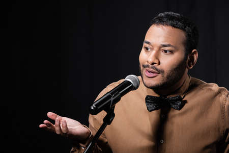 confused eastern stand up comedian telling jokes into microphone on stand isolated on black