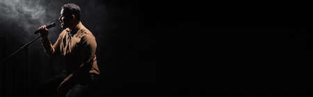 side view of indian comedian sitting on chair and performing stand up comedy into microphone on black with smoke, bannerの写真素材