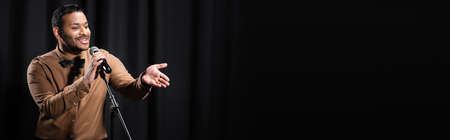 indian comedian performing stand up comedy and telling jokes in microphone while gesturing on black, bannerの写真素材