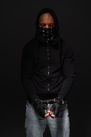 African american bandit with mask on face and handcuffs standing isolated on black