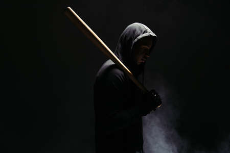 Side view of african american bandit in hood holding wooden baseball bat on black background with...