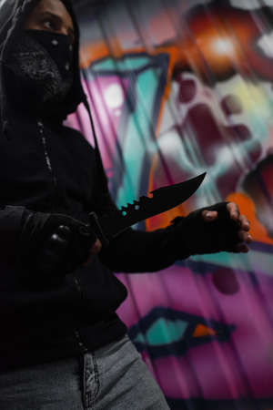 Low angle view of african american bandit in mask holding knife near graffiti outdoorsの写真素材