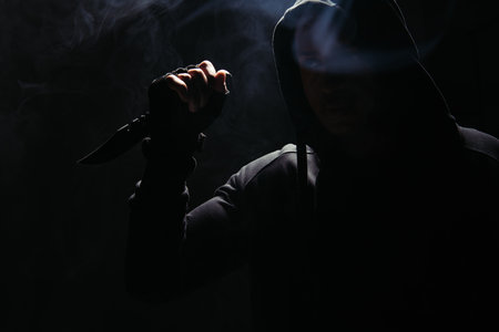 Silhouette of african american bandit in hood holding knife on black background with smokeの写真素材