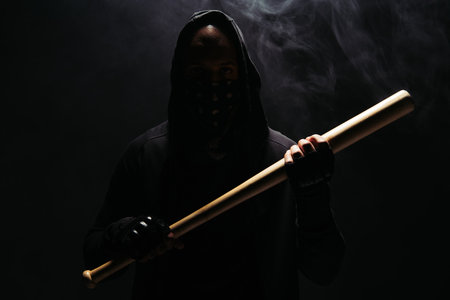 Silhouette of african american man in hoodie and gloves holding baseball bat on black background with smokeの写真素材