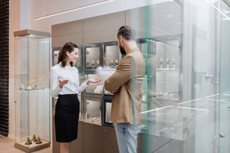 Smiling seller pointing with hands near showcases and customer in jewelry shopの写真素材