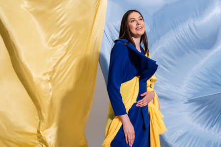 joyful young ukrainian woman in color block clothing posing near blue and yellow fabricの写真素材