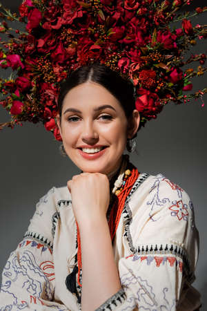 portrait of smiling ukrainian woman in traditional clothes and red wreath with flowers and berries isolated on greyの写真素材