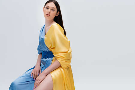 sensual young ukrainian woman in blue and yellow dress posing while sitting isolated on greyの写真素材
