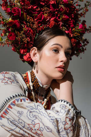 portrait of dreamy ukrainian woman in traditional clothes and floral red wreath isolated on grey