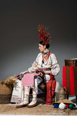 full length of ukrainian woman in traditional clothes and red wreath with flowers holding knitting needles on dark greyの写真素材