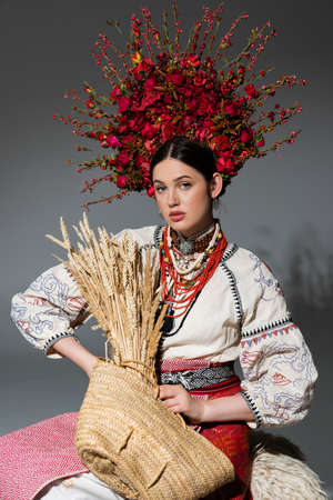 pretty ukrainian woman in traditional clothes and red wreath with berries holding bag with wheat on greyの写真素材