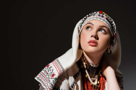 portrait of young ukrainian woman in traditional clothing with ornament and red beads looking up on blackの写真素材