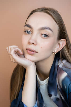 portrait of young and stylish woman with hand near chin and natural makeup isolated on beige