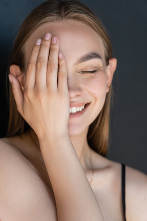 portrait of smiling woman with natural makeup obscuring face with hand isolated on blackの写真素材