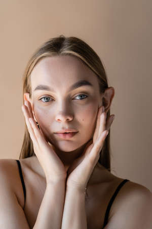 portrait of woman with natural makeup touching face with perfect skin isolated on beigeの写真素材