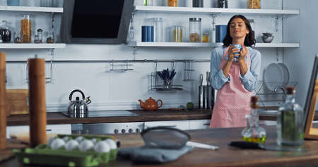 pleased housewife with cup of tea near worktop with food and cooking utensils on blurred foreground