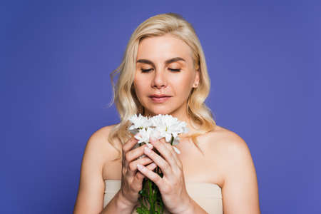 blonde woman with bare shoulders looking at white flowers isolated on violetの写真素材