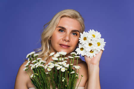 blonde woman with bare shoulders holding white flowers and looking at camera isolated on violet