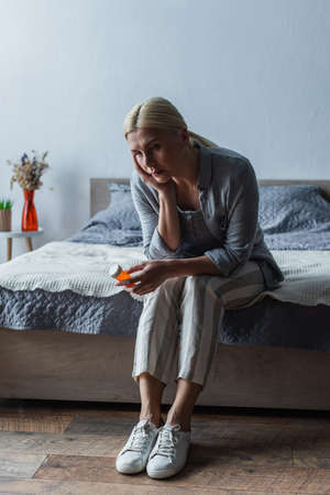 upset blonde woman with menopause holding bottle with pills and sitting on bedの写真素材