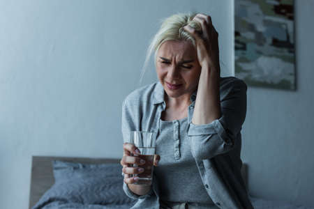 exhausted blonde woman holding glass of water while having migraine during menopauseの写真素材