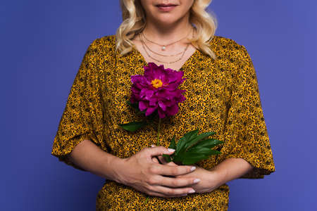 cropped view of blonde woman in blouse holding purple flower with green leaves isolated on violet