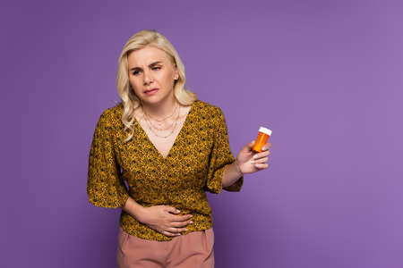 blonde woman with stomach ache holding bottle with pills isolated on purple