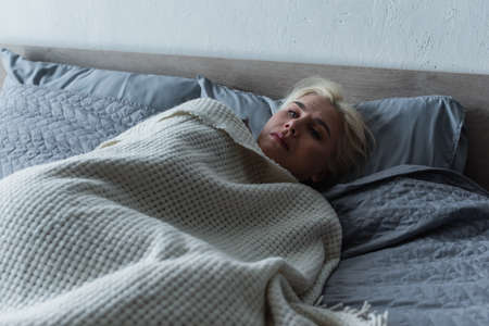depressed blonde woman with menopause lying under blanket in bedの写真素材