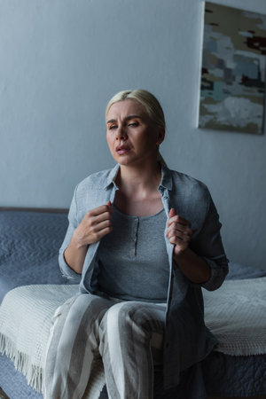 blonde woman with menopause suffering from heat and touching cardigan in bedroomの写真素材