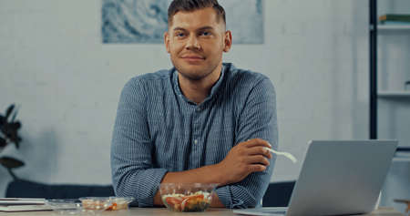 happy man holding plastic fork near salad in container and laptop on desk